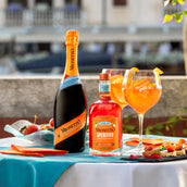 Bottle of champagne, bottle of Cointreau, and two glasses with orange cocktails on a table outdoors.