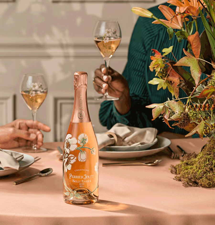 Bottle of Perrier-Jouèt Rosé champagne on a table with people holding glasses and a floral arrangement.