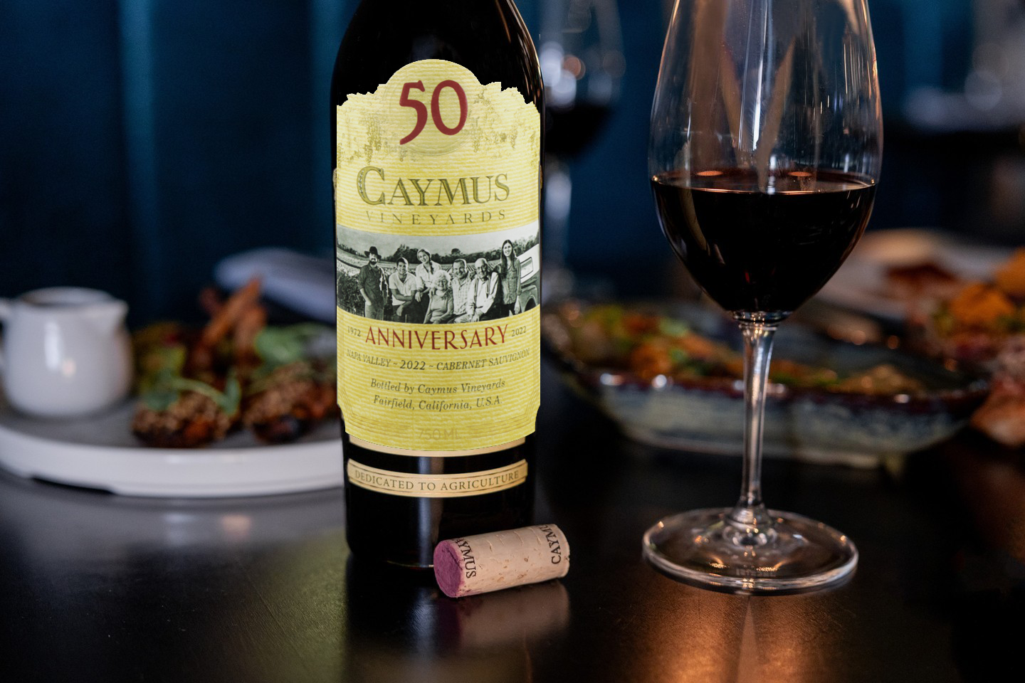 Caymus 50th Anniversary wine bottle with a glass of red wine on a table.