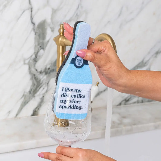 Person holding a novelty wine bottle-shaped dish scrubber with text on a marble background