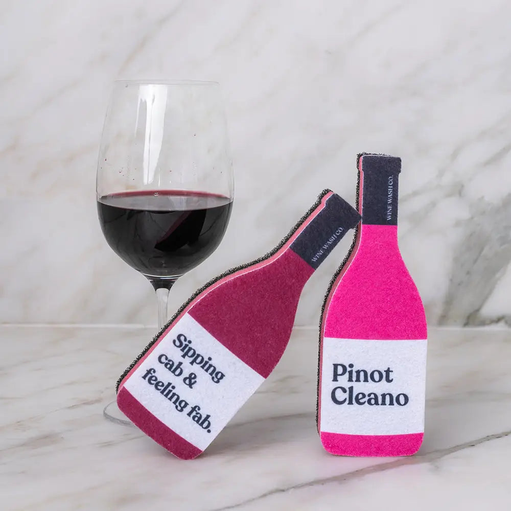 Two pink wine bottle-shaped coasters with text next to a glass of red wine on a marble surface.